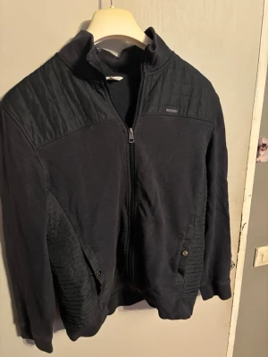 Svart quiltad cardiganjacka - Snygg svart cardiganjacka med quiltade detaljer på axlar och sidor. Jackan har dragkedja framtill, ståkrage och två fickor med knappstängning. Materialet är en mix av mjukt tyg och quiltat tyg som ger en cool kontrast. Perfekt för lager på lager och streetwear.