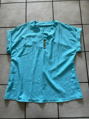 Turkos blus  med knappar storlek 1XL - Säljer en turkos henley med korta ärmar och fyra bruna knappar framtill. Skjortan har en avslappnad passform och är tillverkad i ett lätt bomullsmaterial som känns skönt mot huden. Perfekt för dig som gillar en enkel och stilren look.