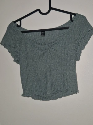 Croptop - Trendig grå croppad topp med ribbad struktur och korta ärmar. Toppen har en lätt rynkad detalj framtill och vågiga kanter vid ärmslut och nederkant. Perfekt för dig som gillar en avslappnad men snygg stil. Aldrig använd
