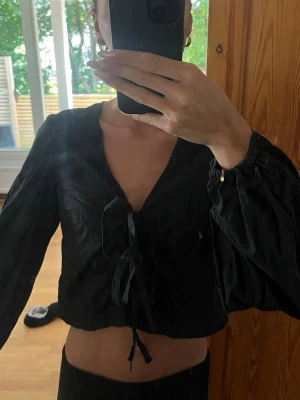 Svart omlott blus med snörning - Svart blus med omlott-design och snörning framtill. Blusen har långärmade ärmar och en croppad passform 