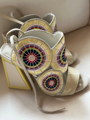 Unika pumps från Kat Maconie - Säljer ett par statement pumps från Kat Maconie med höga blockklackar och öppen tå. Skorna är beige med färgglada broderade cirkelmönster i gult, blått, rött och svart. De har snörning och detaljer i lackat material. Perfekta för dig som vill sticka ut!