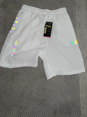 Vita träningsshorts från Under Armour - Snygga vita shorts från Under Armour med elastisk midja och snörning. Loggan och texten ARMOUR syns på sidan i regnbågsskimrande och svart tryck. Perfekta för träning eller chill. Lätta och bekväma i syntetmaterial.