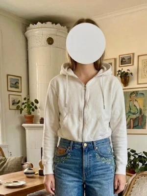 Vit hoodie med dragkedja och huva - Klassisk vit hoodie med dragkedja framtill, huva med dragsko och två fickor. Mjuk bomullskänsla och ribbade muddar vid ärmslut och nederkant. Perfekt för en avslappnad stil.