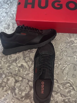 Svarta sneakers från HUGO - Använda 2-3 gånger nypris 3,2 Stilrena svarta sneakers från HUGO med röda logodetaljer. Skorna har en klassisk låg siluett, ovandel i en mix av läder och textil samt svarta snören. Perfekta för en clean och modern look