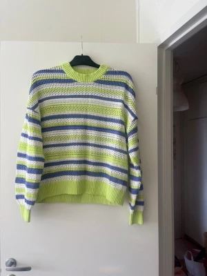 Randig stickad tröja i lime och blått - Snygg stickad tröja med breda ränder i limegrönt, blått och vitt. Tröjan har rund halsringning, ribbade muddar och en luftig stickning som ger en cool look. Perfekt för dig som gillar färg och vill sticka ut lite extra.