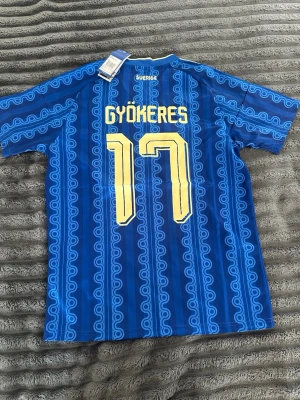 Blå Sverige fotbollströja Gyökeres 17 - Säljer en blå Sverige fotbollströja från Adidas med Gyökeres och nummer 17 på ryggen. Tröjan har gula detaljer, klassiskt mönster och Sveriges landslagsemblem på bröstet. Perfekt för dig som vill stötta landslaget med stil.