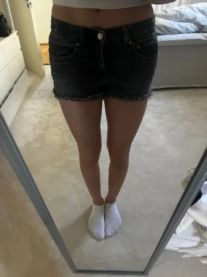 Svarta jeansshorts med fransig kant - Säljer ett par svarta lågmidjade jeansshorts med fransig, rå kant och klassisk femficksdesign. Shortsen har knapp och dragkedja framtill samt midwaist passform. Perfekta för sommaren och ger en avslappnad vibe.