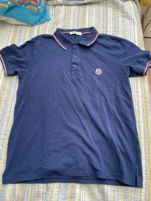 Marinblå pikétröja från Moncler - Klassisk marinblå pikétröja från Moncler med krage och knappar framtill. Tröjan har röda och vita ränder på kragen och ärmsluten samt Moncler-logga broderad på bröstet. Perfekt för en stilren och sportig look.