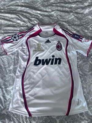 AC Milan Champions League tröja - Vit AC Milan matchtröja från Adidas med röda och svarta detaljer, Champions League-märken på ärmarna och klubbmärke på bröstet. Tryckt sponsor 'bwin' på framsidan. Tillverkad i funktionsmaterial, perfekt för fotbollsträning eller support.
