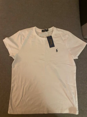 Vit t-shirt från Polo Ralph Lauren - Klassisk vit t-shirt från Polo Ralph Lauren med korta ärmar och rund hals. T-shirten har en liten broderad logga i mörkblått på bröstet och är gjord i mjuk bomull. Perfekt basic-plagg med snygg passform.