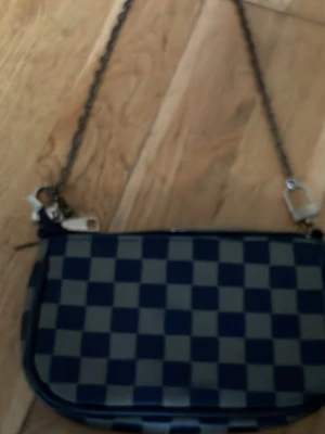 Rutigt axelremsväska i blått och grått - Snygg handväska med blå och grå rutigt mönster och kedjerem. Väskan har en rektangulär form och dragkedja upptill. Materialet ser ut att vara konstläder eller canvas. Perfekt för dig som gillar klassisk men trendig stil.