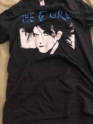 Svart The Cure t-shirt med tryck - Cool svart t-shirt med The Cure-tryck i blått och vitt framtill. Klassisk passform med rund hals och korta ärmar. I bra skick borsett från storlekslappen.