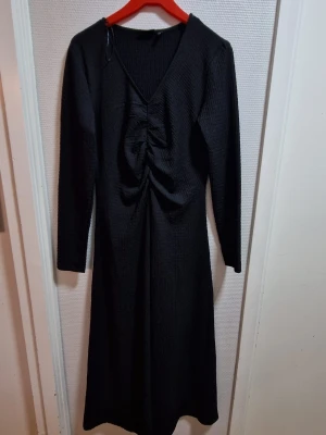 Svart långärmad klänning med v-ringning - Svart långklänning med v-ringad hals och rynkade detaljer framtill. Klänningen har lång ärm och är tillverkad i ett stretchigt material med diskret struktur. Perfekt för dig som gillar stilrena och enkla plagg med en twist.