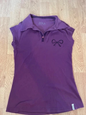 Lila pikétopp med rosettdetalj Zola - Snygg lila pikétopp från Zola med korta ärmar och klassisk krage. Toppen har en liten rosettdetalj broderad på bröstet och knäppning framtill. Perfekt för en trendig och avslappnad look.