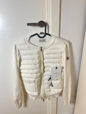 Vit cardiganjacka från Moncler - Stilren vit cardiganjacka från Moncler med quiltad front, två fickor med lock och dragkedja. Jackan har rund halsringning, ribbade muddar och Moncler-logga på ärmen. Materialmix av stickat tyg och vadderad front ger en modern look.