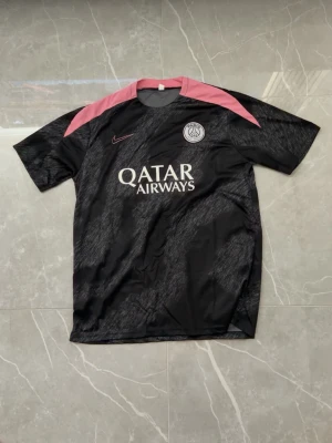 PSG svart och rosa matchtröja Nike - Snygg svart PSG matchtröja från Nike med rosa detaljer på axlarna och diskret mönster över hela tröjan. Tryck med Qatar Airways på bröstet och PSG-logga på vänster sida. Perfekt för fotbollsträning eller att bära som supporter.