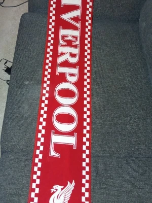 Röd Liverpool halsduk - Röd halsduk med stora vita bokstäver där det står 'LIVERPOOL' och klubbens logga. Klassisk supporterhalsduk med rutiga kanter i vitt och rött, perfekt för att visa ditt stöd på matchen.