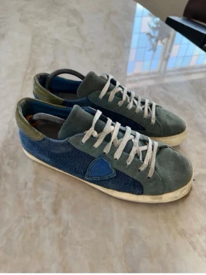Blå och gröna sneakers från Philippe Model - Snygga sneakers från Philippe Model i blått och grönt med ovandel i mocka och textil. Skorna har vita snören, blå detalj på sidan och en klassisk låg siluett. Perfekta för dig som gillar en unik och stilren look.