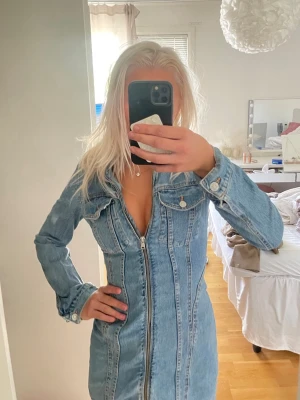 Ljusblå jeansklänning med dragkedja - Snygg ljusblå jeansklänning med lång ärm och hel dragkedja framtill. Klänningen har markerade sömmar och bröstfickor med knappdetaljer. Perfekt för dig som gillar en cool och trendig stil.