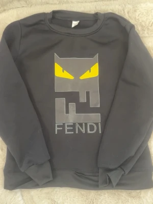 Svart Fendi sweatshirt med tryck - Cool svart sweatshirt från Fendi med ett stort grafiskt tryck framtill som har gula ögon och Fendi-loggan. Tröjan har rund halsringning, långa ärmar och ribbade muddar. Perfekt för dig som gillar streetwear och vill sticka ut med en unik design.