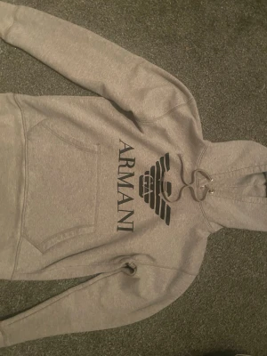 Grå hoodie från Armani med logga - Snygg grå hoodie från Armani med stor svart logga och text på bröstet. Hoodien har huva med dragsko och en stor magficka framtill. Perfekt för en chill och stilren look. Mjuk och skön bomullsblandning.