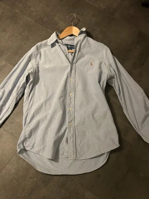 Ljusblå skjorta från Ralph Lauren - Klassisk ljusblå skjorta från Ralph Lauren med knappar framtill och broderad logga på bröstet. Skjortan har lång ärm, krage och är tillverkad i bomull. Perfekt för sommaren & en stilren och avslappnad look.