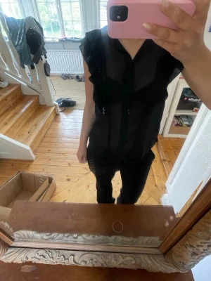Svart transparent blus med volang - Snygg svart blus i transparent material med knäppning framtill och volangdetaljer vid axlarna. Blusen har korta ärmar och en lätt, luftig känsla som passar perfekt till jeans eller kjol. 