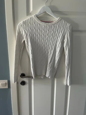 Vit kabelstickad tröja - Snygg vit kabelstickad tröja med rund halsringning och långa ärmar. Tröjan har ett klassiskt flätmönster och ribbade muddar vid ärmslut och nederkant. Perfekt att matcha med jeans eller kjol för en stilren look. 134/140
