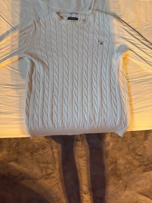 Vit kabelstickad tröja från Gant - Kabelstickad vit tröja från Gant med rund halsringning och diskret logga broderad på bröstet. Tröjan har långa ärmar och ribbade muddar vid ärmslut och nederkant. Perfekt för lager-på-lager och klassisk stil.