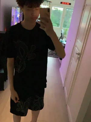 Syna World  - Säljer ett svart set med t-shirt och shorts, båda med stora graffiti-inspirerade tryck i grått. T-shirten har rund hals och korta ärmar, shortsen har resår i midjan. Perfekt för dig som gillar streetwear och en avslappnad stil.