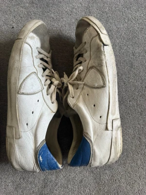 2 Vita sneakers med blå detalj, oliver Cadell, Philippe model - Säljer ett par vita sneakers med blå häl och gråa mockadetaljer. Skorna har en låg profil, vita snören och en unik cirkeldesign på sidan. Materialet är en mix av läder och mocka. Perfekta för dig som gillar en clean men ändå cool look. Fin för Sommar.Priset kan diskuteras.