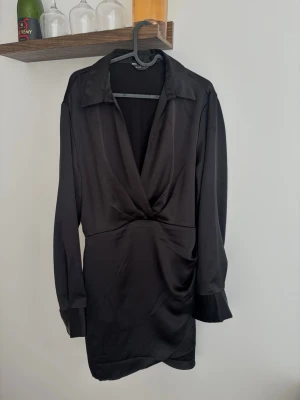 Svart omlottklänning i satin - Stilren svart kort omlottklänning i satin med lång ärm och djup v-ringning. Klänningen har en lätt draperad effekt vid midjan och klassisk krage, perfekt för en trendig look.