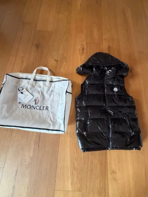 Svart dunväst från Moncler med huva - Snygg svart dunväst från Moncler med glansig finish och avtagbar huva. Västen har Moncler-logga på bröstet och praktiska fickor framtill. Perfekt för lager-på-lager och streetstyle. Kommer med originalpåse.