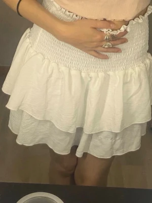 Vit volangkjol med smock - Supersöt vit kortkjol med dubbla volanger och bred smock i midjan. Kjolen har en luftig och flowy känsla, perfekt för varma dagar. Passar till crop tops och sneakers för en chill sommarlook.