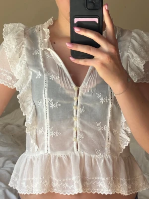 Vit broderad blus med volanger - Romantisk vit blus i tunn bomull med broderade blommor, volangdetaljer längs ärmar och knappslå framtill. Kortärmad modell med v-ringning och söt spetskant nedtill.  