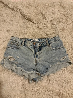 Ljusa jeansshorts från ZARA - Snygga ljusblå jeansshorts från ZARA med slitna detaljer och fransig kant. Klassisk femficksmodell med hög midja och dragkedja. Perfekta för sommaren