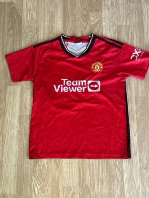 Manchester United Rashford matchtröja - Röd Manchester United matchtröja med svarta och vita detaljer, officiellt klubbmärke på bröstet och TeamViewer-logga. På ryggen står det Rashford och nummer 10. Perfekt för fotbollsfans som vill visa sitt stöd på läktaren eller planen.