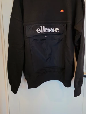 Svart sweatshirt från Ellesse - Svart sweatshirt från Ellesse med stor ficka framtill och tydlig logga i vitt. Tröjan har rund halsringning och långa ärmar. Materialet är mjukt och bekvämt, perfekt för en avslappnad stil.