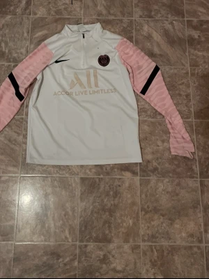 PSG träningströja med rosa ärmar - Snygg långärmad träningströja från Nike för PSG med vita och ljusrosa färger. Tröjan har halv dragkedja, svarta detaljer på ärmarna och PSG-logga på bröstet. Materialet är lätt och andas, perfekt för fotbollsträning eller sport.