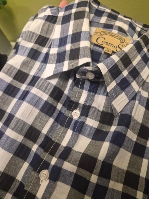 Blårutig kortärmad skjorta CosmoS XL - Kortärmad skjorta från CosmoShirt i blått och vitt rutigt mönster. Skjortan har klassisk krage, knäppning framtill med vita knappar och en bröstficka. Perfekt för en avslappnad och stilren look.