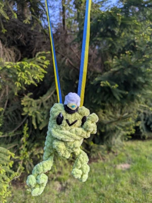 Groda som studenthänge 🐸 - En söt handgjord groda i ljusgrönt garn med svart broderade ögon och ett litet studentmössa på huvudet. Hänger i ett blågult band, perfekt som dekoration eller present vid studentfirande.