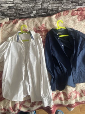 Vit och marinblå skjorta herr - Två klassiska skjortor, en vit och en marinblå. Båda har krage och knäppning framtill, samt långa ärmar. Den vita skjortan har en liten broderad logga på bröstet och kontrastfärgad insida på kragen. Perfekta för en stilren look.