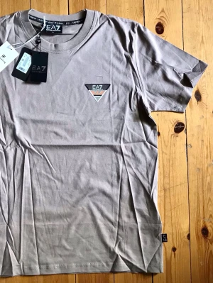 Beige EA7 t-shirt med logga - Snygg beige t-shirt från EA7 Emporio Armani med rund hals och korta ärmar. T-shirten har en liten EA7-logga på bröstet och är gjord i mjuk bomull. Perfekt för en stilren och avslappnad look.