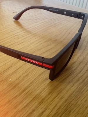 Svarta solglasögon från Prada - Snygga svarta solglasögon från Prada med rektangulära bågar och diskret logga på sidan. Bågarna är i matt svart plast med en röd detalj och Prada-text på skalmen. Perfekt accessoar för en trendig look.