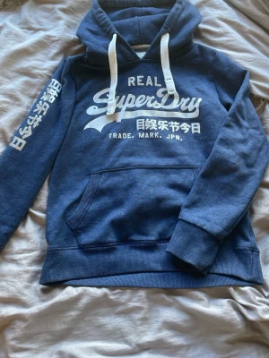 Blå Superdry hoodie med tryck - Säljer en blå hoodie från Superdry med vit logga och japansk text på bröstet och ärmen. Hoodien har en stor ficka framtill, vita dragsnören och ribbade muddar. Perfekt för dig som gillar streetwear och vill ha något bekvämt och snyggt.