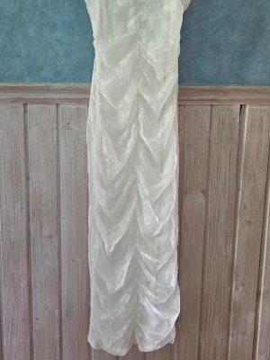 Vit spetsklänning lång modell - Elegant långklänning i vit spets med ärmlös design. Klänningen har ett vackert spetsmönster och draperad effekt framtill som ger en lyxig känsla. Perfekt för speciella tillfällen där du vill sticka ut med en klassisk och stilren look.