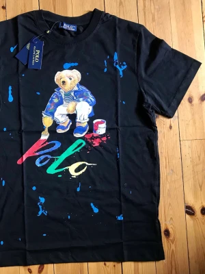 Svart Polo Ralph Lauren t-shirt med björn - Cool svart t-shirt från Polo Ralph Lauren med färgglad björn- och logotyptryck framtill. T-shirten har korta ärmar och rund halsringning. Materialet är mjuk bomull och trycket har stänk i flera färger för en kreativ look.