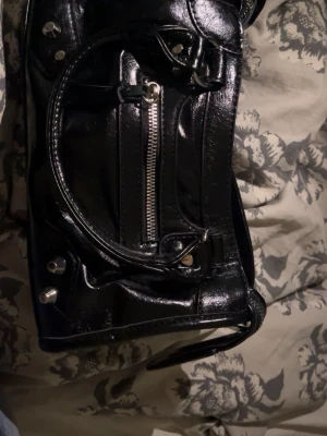 Svart handväska med silverdetaljer - Snygg svart handväska i glansigt material med silverfärgade metalldetaljer och dragkedjor. Väskan har dubbla handtag, dekorativa spännen och en framficka med dragkedja. Perfekt för dig som gillar en edgy och trendig look.