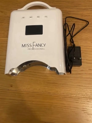 Miss Fancy UV-lampa för naglar - UV-lampa från Miss Fancy Professional i vit plast med digital display och timerknappar. Perfekt för att härda gelélack och andra nagelprodukter. Lampan har en praktisk bärhandtag och medföljer strömadapter. Modern design med silverfärgad insida.