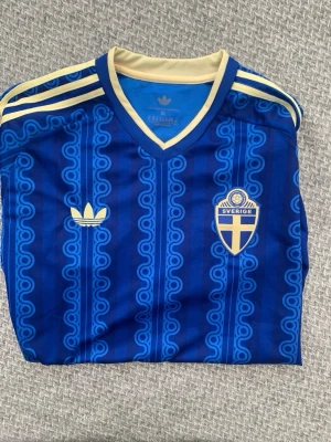 Blå Sverige fotbollströja Adidas XL - Säljer en blå fotbollströja från Adidas i storlek XL med Sveriges landslagsemblem på bröstet. Tröjan har retroinspirerat mönster, V-ringning och gula detaljer på axlar och krage. Perfekt för dig som vill stötta Sverige med stil.